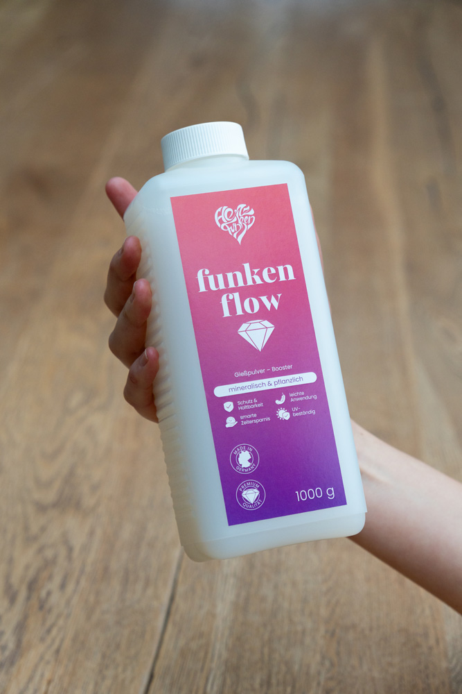 Eine Hand hält die 1000 g Flasche Funkenflow Gießpulver-Booster vor einem Holzhintergrund.